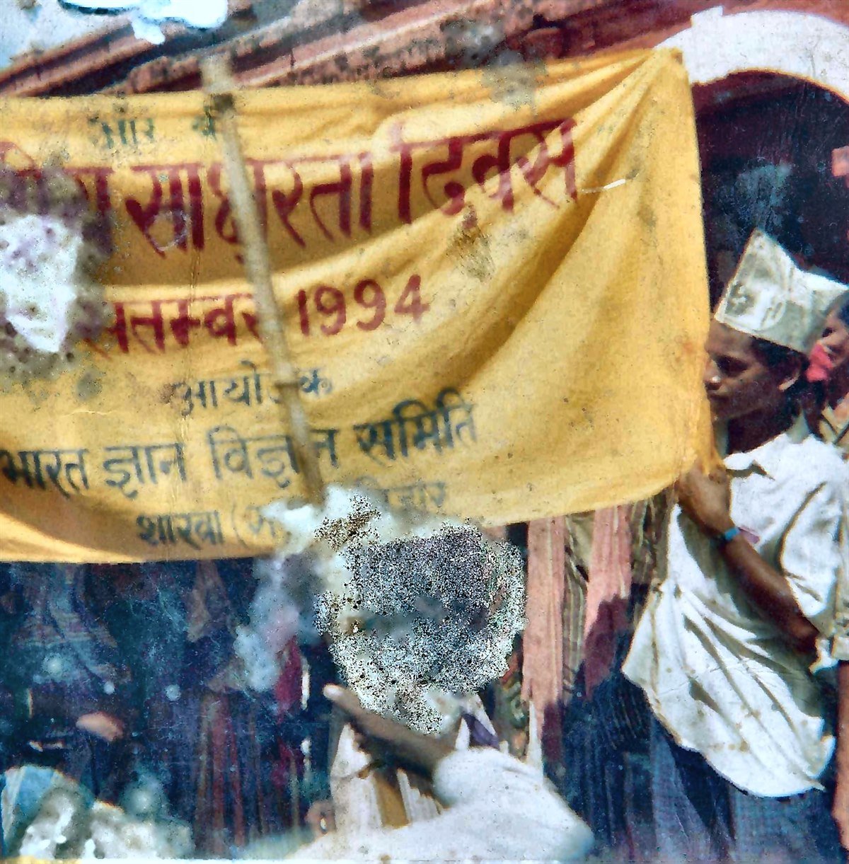 A restored 1994 photograph: volunteers in paper caps carry a yellow banner reading 'राष्ट्रीय साक्षरता दिवस · सितम्बर 1994 · आयोजक: भारत ज्ञान विज्ञान समिति'.
