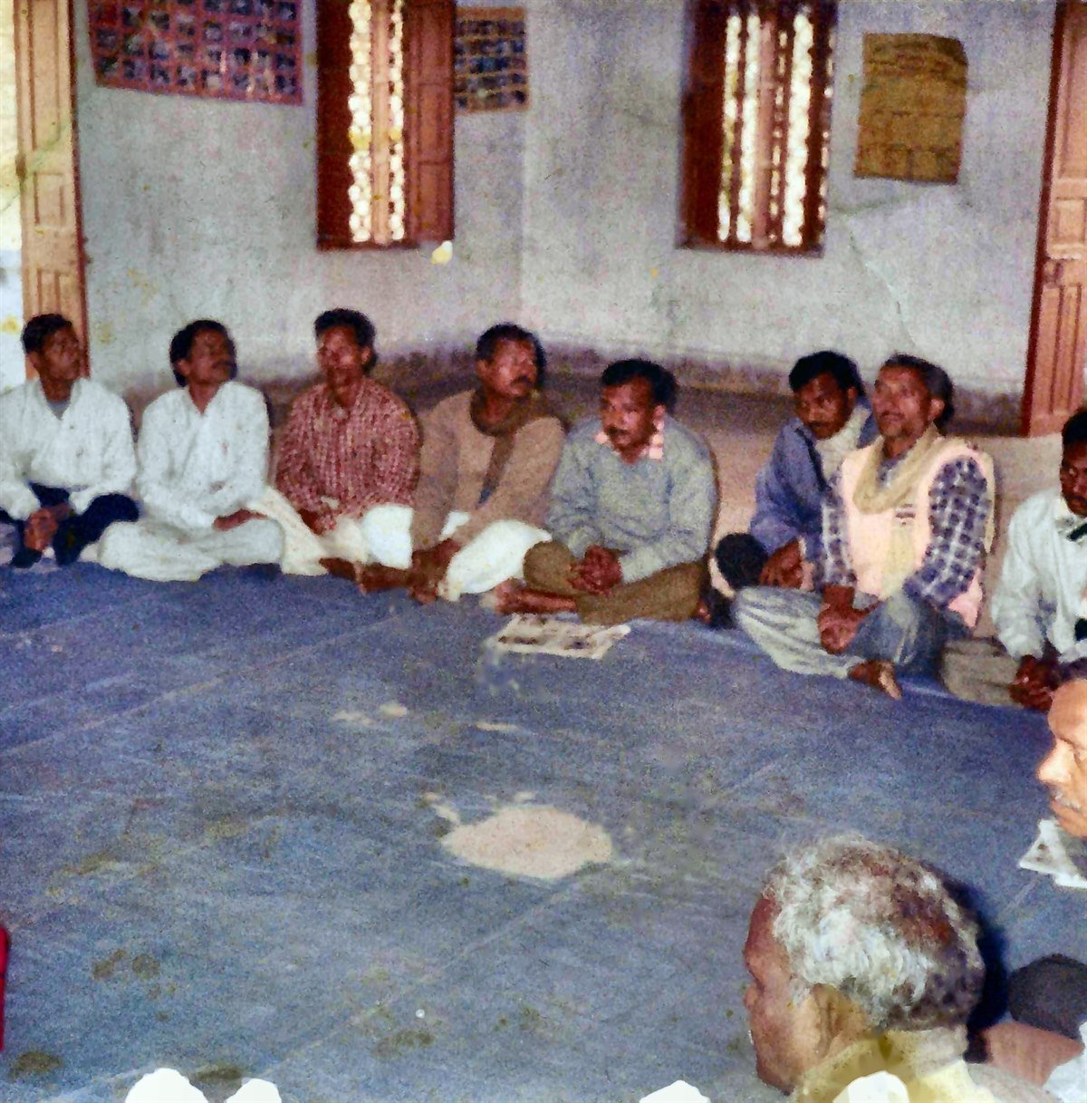 Restored archive photograph of about twenty members seated in a circle at a samiti meeting, with a lamp on a draped table.
