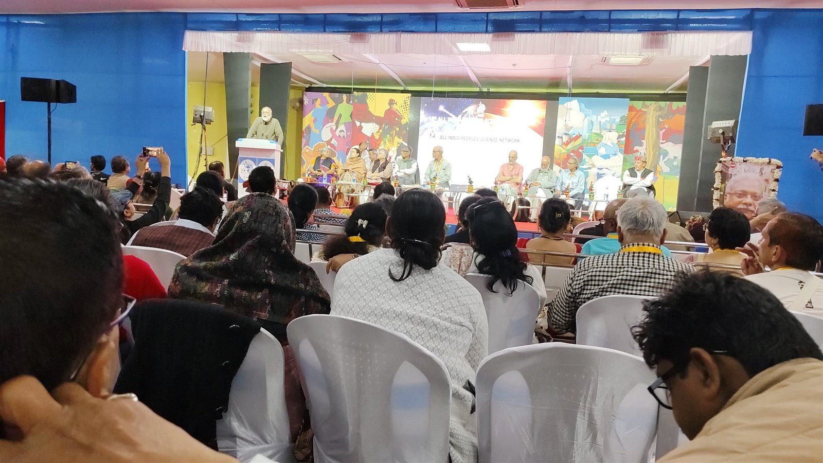 A packed hall at an All India People's Science Network national gathering, with a panel on stage and the AIPSN name on the screen behind, December 2024.