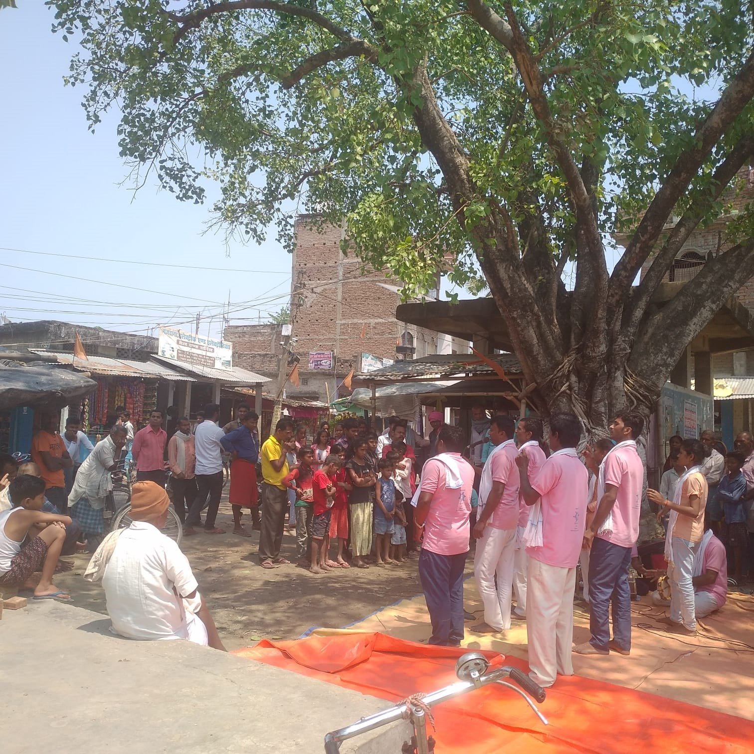 A Kala Jatha troupe in matching pink kurtas performing under a peepal tree in a village square at Gaya, with villagers watching and a dholak on the ground, April 2023.