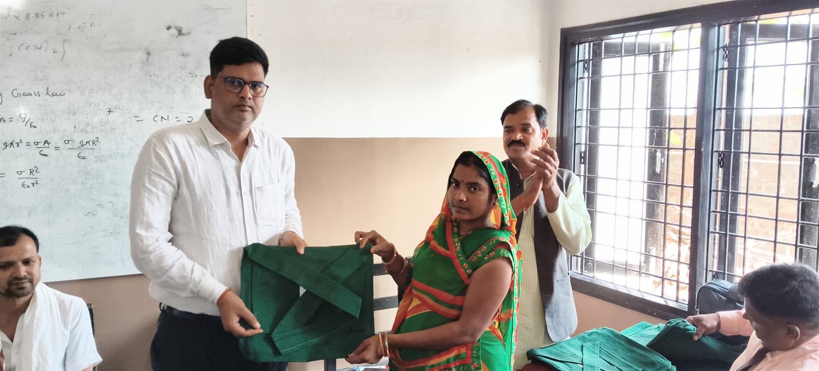 A samiti worker hands a bag of learning material to a woman in a classroom, May 2026.