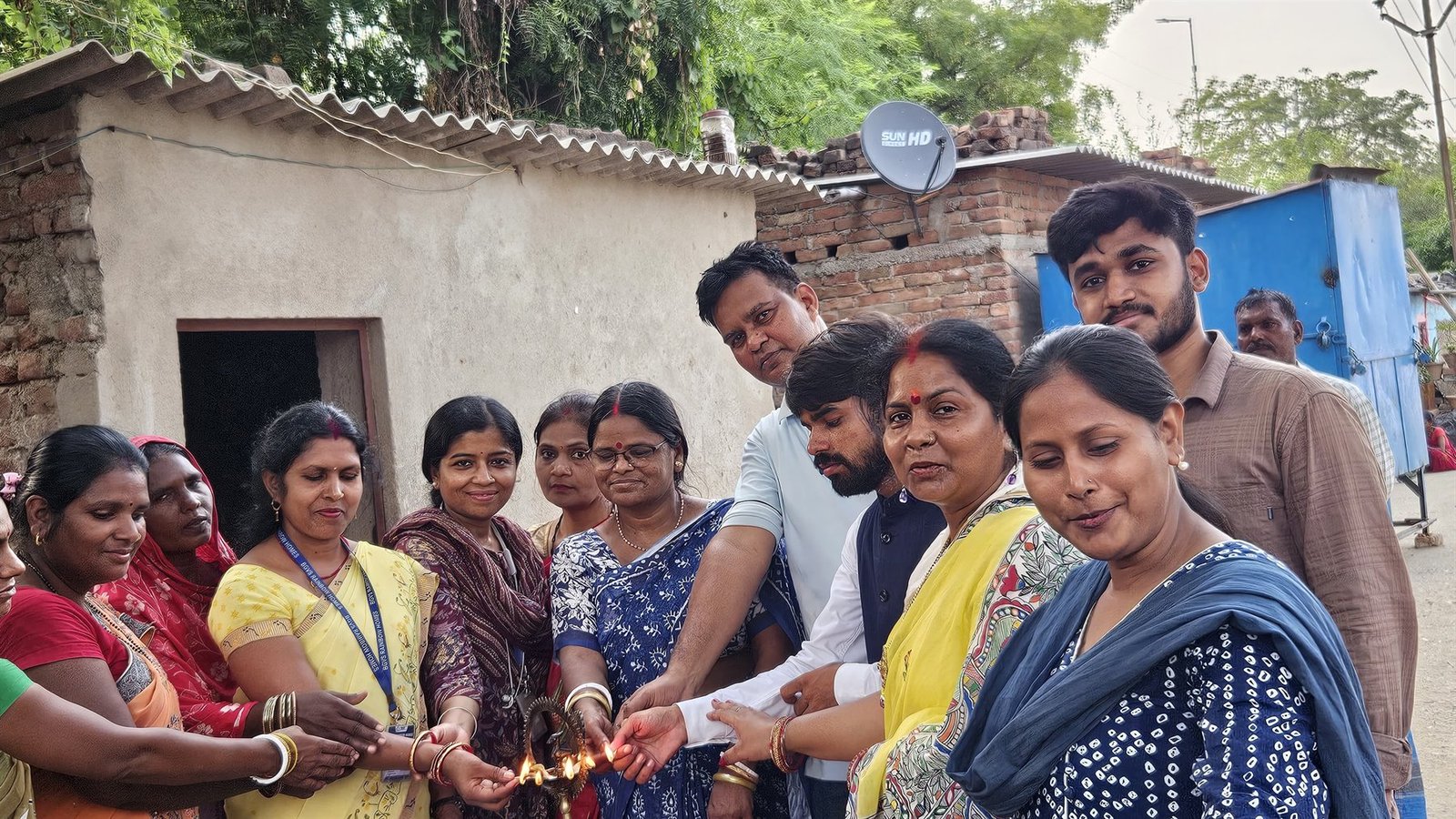 Women of the community and samiti workers lighting an oil lamp together outside a Rainbow Home in June 2026, wearing BGVS Rainbow Homes lanyards.