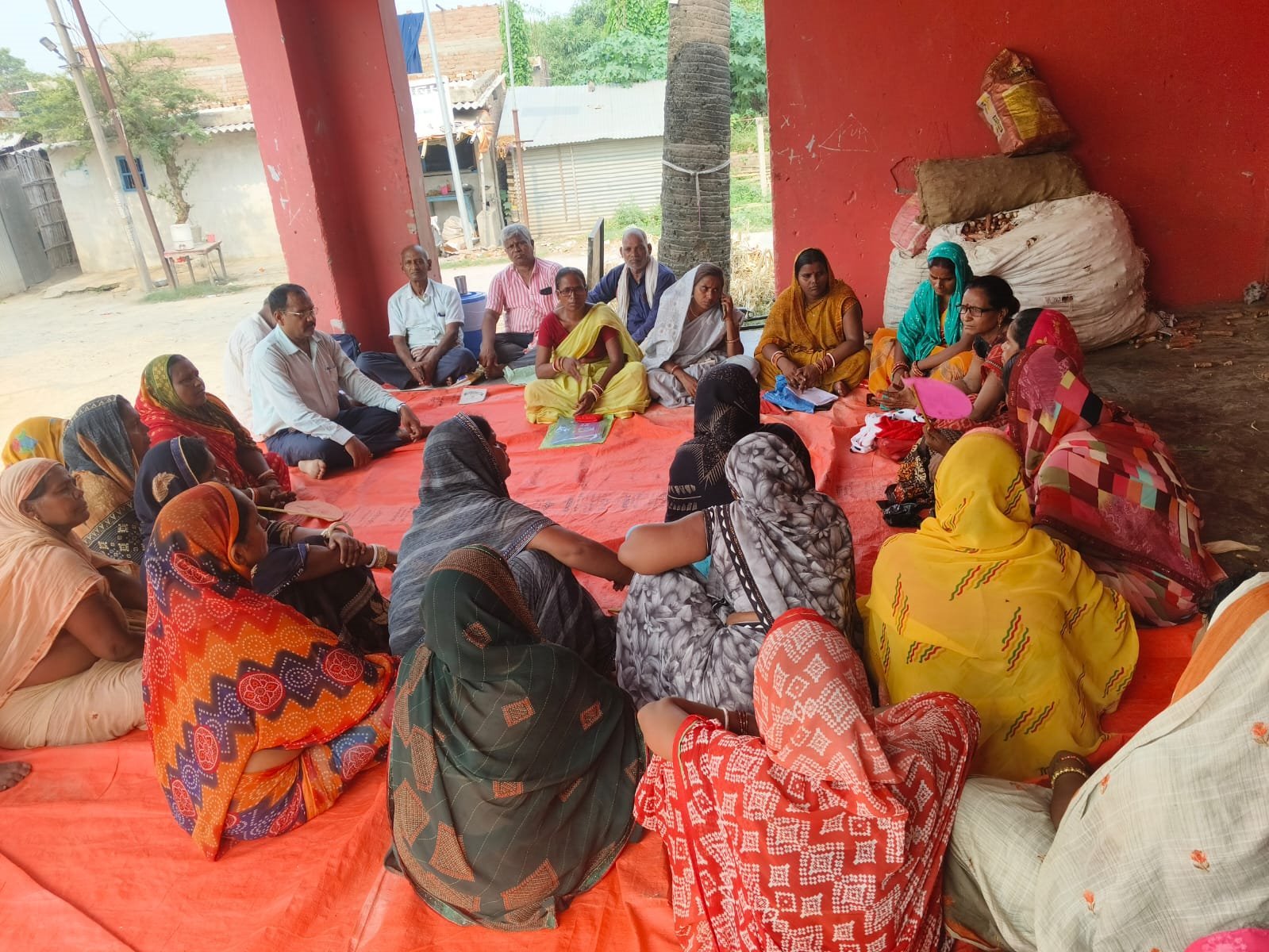 Members of a village Buniyadi Sabha seated in a circle on an orange mat, mostly women in bright saris, Begusarai district, June 2026.