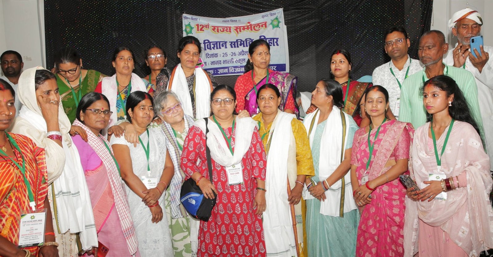A large group of women delegates wearing pass cards, standing together beneath the banner of the 12th State Conference of Gyan Vigyan Samiti, Bihar, at Chand, Kaimur, April 2026.